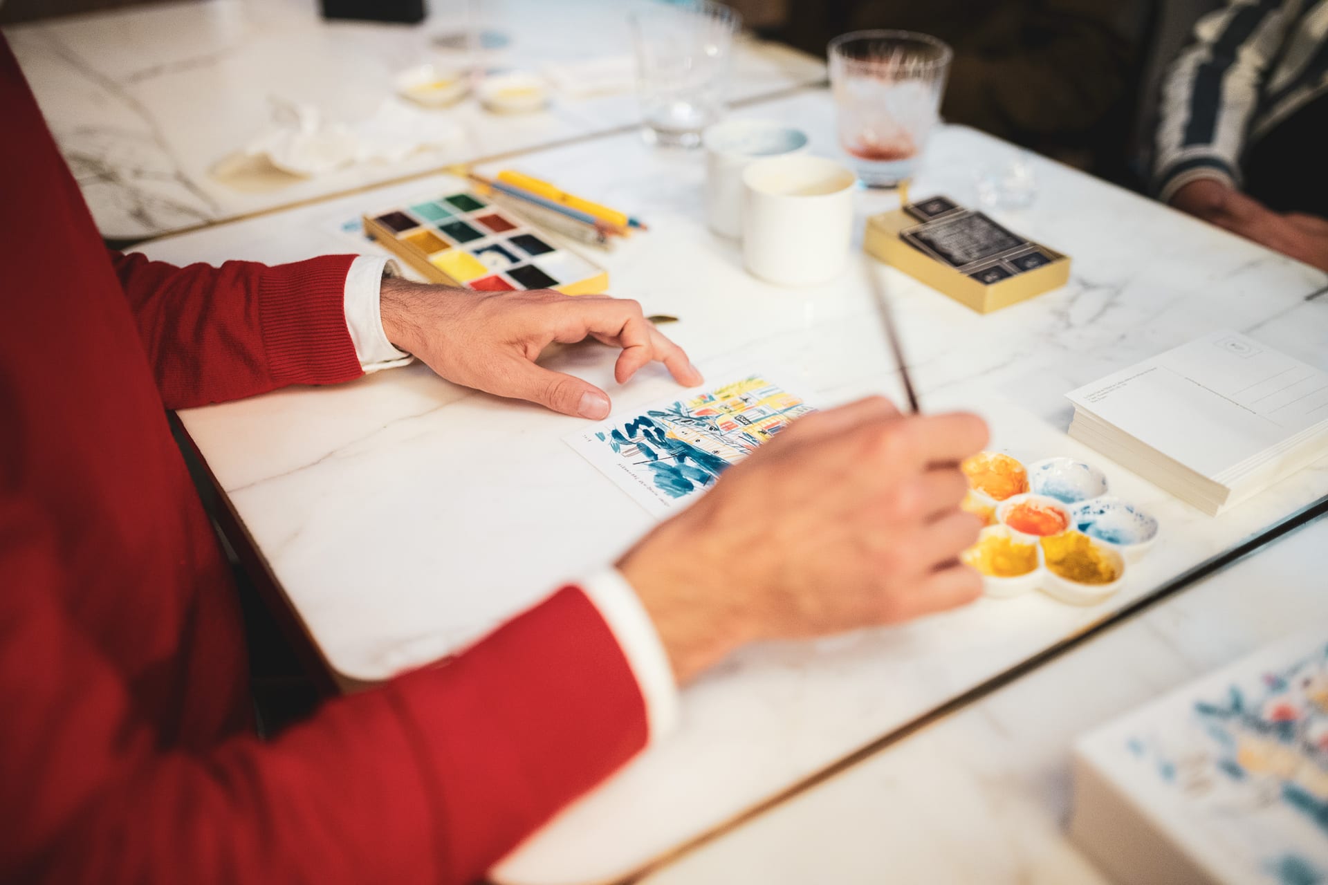 Un homme en pull rouge peint une carte sur une table avec des aquarelles.